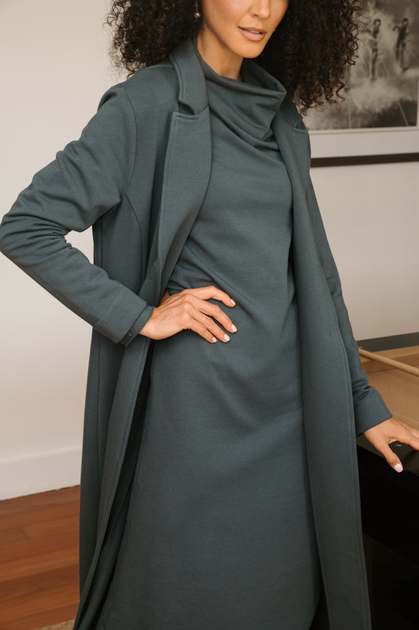 Woman wearing a long grey coat over a dress indoors.