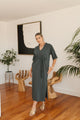 Woman in a gray dress standing in a modern interior setting with plants and wooden furniture.