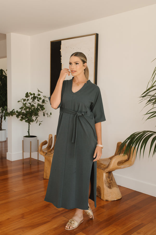 Woman in a grey dress standing in a room with wooden floor and plants.
