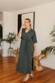 Woman in a grey dress standing in a room with wooden floor and plants.