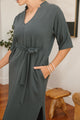 Person wearing a grey dress with a belt in a room with wooden furniture.
