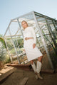Woman in a white dress and knee-high boots standing in front of a greenhouse.