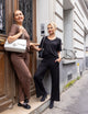 two women, one wearing brown shirt and pants, with white bag over shoulder, the other wearing black shirt and pants, and black bag over shoulder