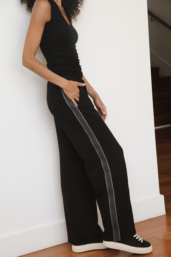 Person wearing a black outfit with a white stripe down the pant leg standing against a white wall.