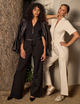 two women, one wearing black shirt, leather jacket with gold zipper and black pants, and the other wearing white shirt and white pants