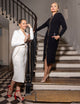 two women, one wearing long white dress with gold zipper and white belt, the other wearing long black dress, with gold zipper and black belt