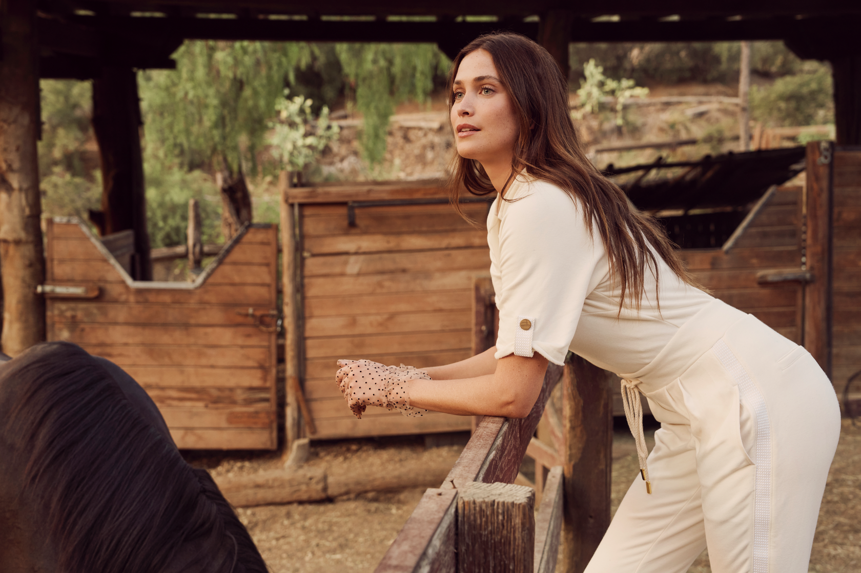 A brown haired woman in a white jumpsuit leans against the fence of a horse paddock.