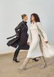 Two women walk past each other. One wears a black dress and trench coat and the other wears a white dress and trench coat