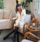 A brown haired woman sits on a white wood and cloth chair. She is wearing a white long sleeve dress and knee-high black leather boots.