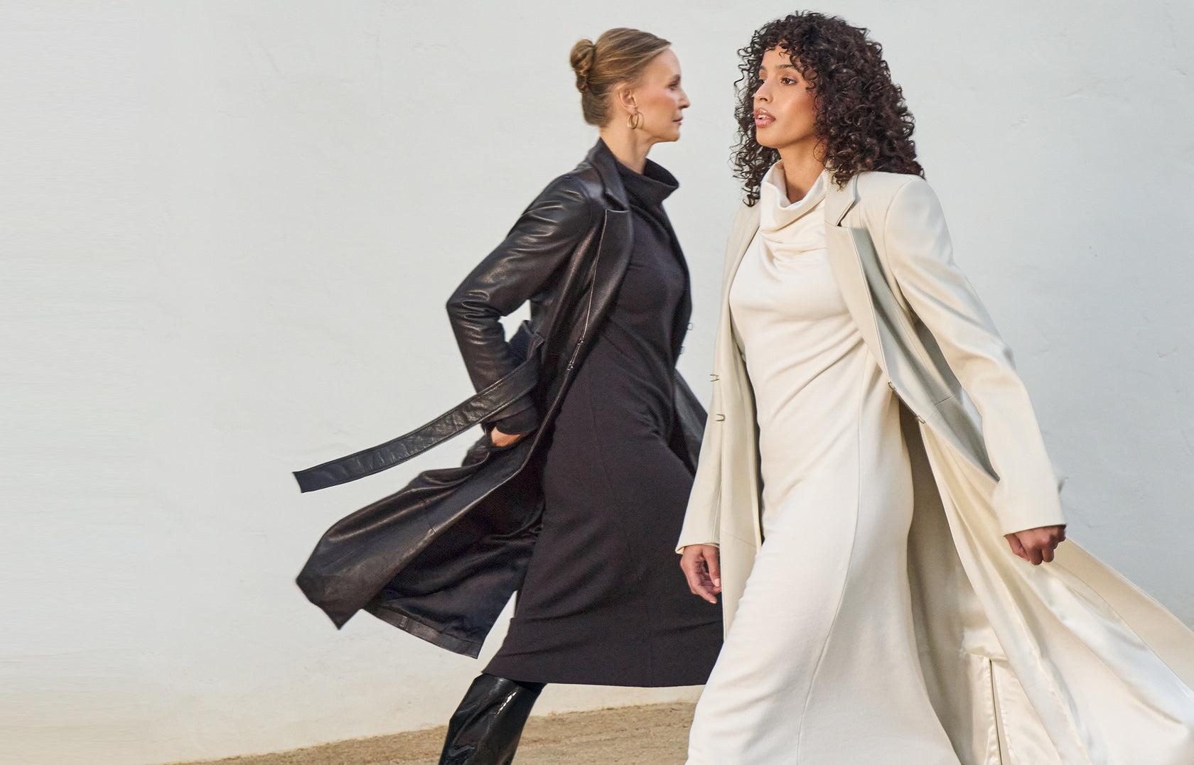 Two women walk past each other. One wears a black dress and trench coat and the other wears a white dress and trench coat