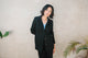 Woman wearing a black blazer over a blue top against a beige wall with plants.