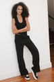 Woman wearing a black sleeveless top and pants with white sneakers against a white wall.