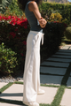 Person wearing a green top and white pants standing on a garden path with plants and flowers in the background.