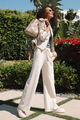 Woman in a stylish outfit with a beige jacket and white pants, holding a beige handbag, standing outdoors.