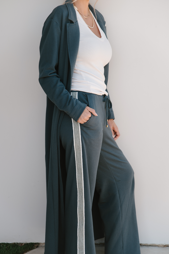Person wearing a long grey coat over a white top and gray pants against a white wall.