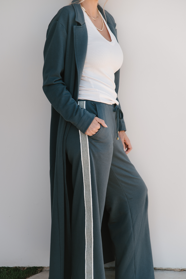Person wearing a long grey coat over a white top and gray pants against a white wall.