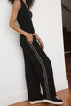 Person wearing a black outfit with a white stripe down the pant leg standing against a white wall.